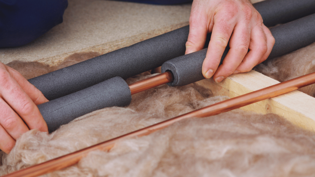 A person installing foam insulation on copper water pipes, as a method for preventing frozen pipes, with thick fiberglass insulation visible in the background.