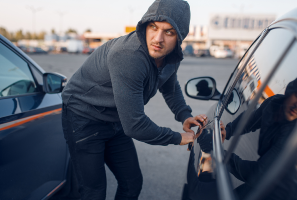 A man in a gray hoodie is attempting to break into a black car with a slim jim tool. He is looking over his shoulder with a cautious expression. There is a clear reflection of his face in the car window.